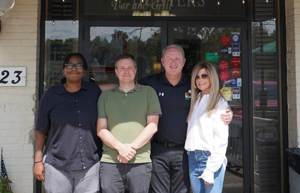 Four adults stand smiling in front of a restaurant entrance, posing together. Two men are in the center, with a woman on each side. Sunlight brightens the scene, and various colorful stickers are on the glass door behind them.