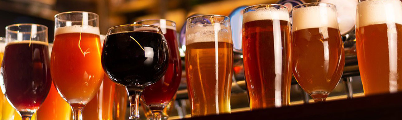 beers-banner A row of various beer glasses, each filled with different types and colors of craft beer, lined up on a bar with blurred lights in the background.