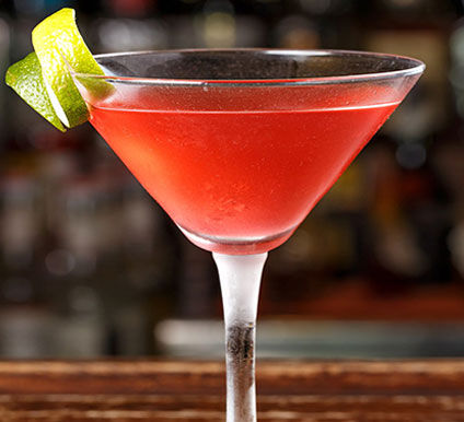 cocktail1 A red cocktail in a martini glass, garnished with a twisted lime peel, sits on a wooden bar with a blurred background.