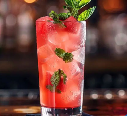 cocktail2 A tall glass filled with a red iced drink, garnished with fresh mint leaves and crushed ice, sits on a dark surface with a blurred background.