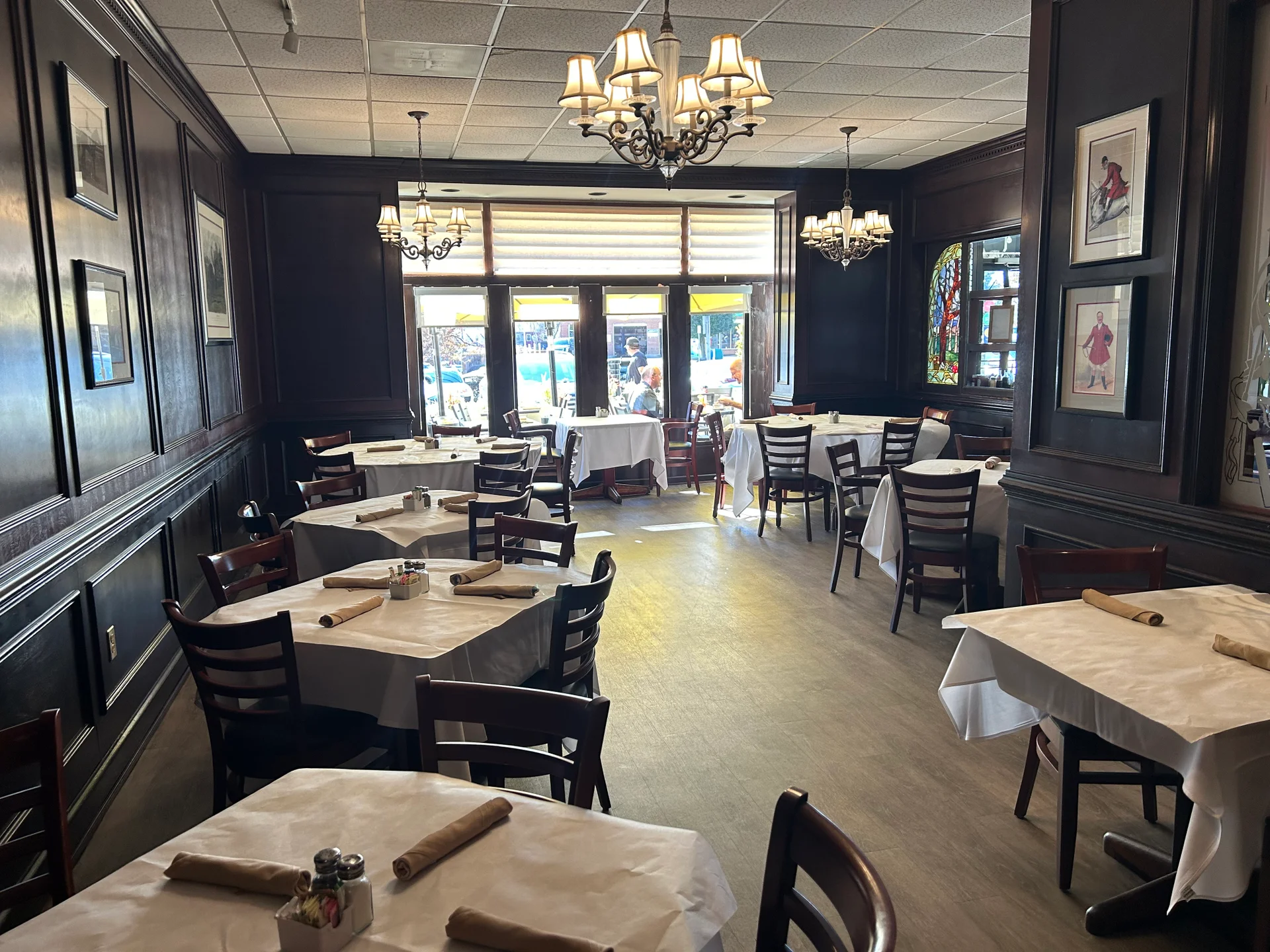 A restaurant dining room with dark wood walls, white tablecloths, wooden chairs, chandeliers, framed art, and large windows letting in sunlight from the street outside. Some tables are set and unoccupied.