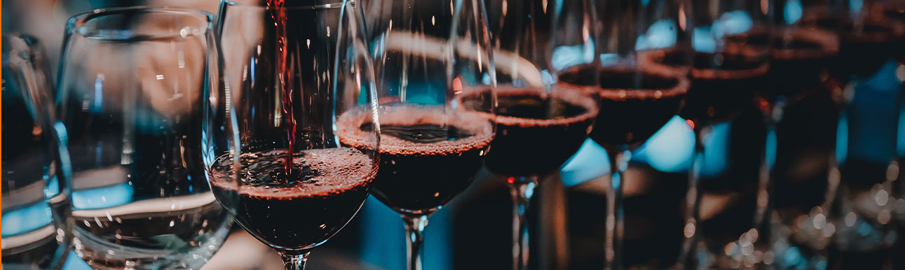 red-wines-drinks A row of wine glasses being filled with red wine, with bubbles forming on the surface, set against a softly blurred background.