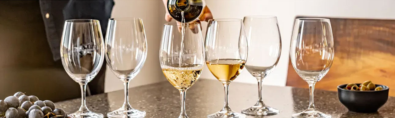 white-wines A hand pours white wine into one of six wine glasses on a countertop, with grapes and a bowl of olives nearby.