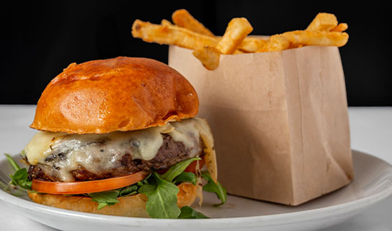 A cheeseburger with lettuce and tomato on a bun sits next to a brown paper bag filled with French fries on a white plate.