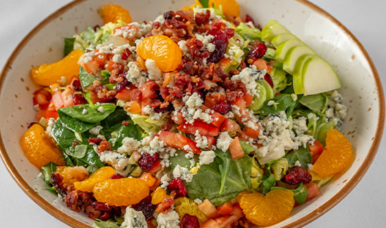 A bowl of salad with spinach, blue cheese crumbles, diced tomatoes, apple slices, mandarin oranges, dried cranberries, and bacon bits.